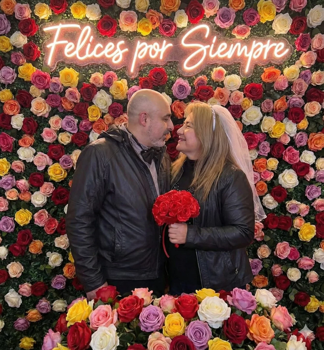 Frank y Nel frente a un muro de flores con el letrero Felices por Siempre